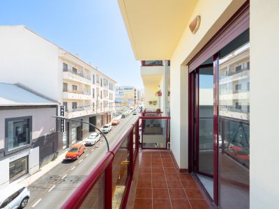 Zonnig balkon met terracotta vloertegels en een rood omrand glazen balustrade, met uitzicht op een stedelijke straat met gebouwen.