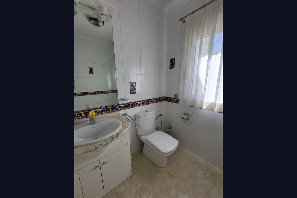 Badkamer met witte tegelwanden en decoratieve rand. Voorzien van een wastafel met licht marmeren blad, een toilet en een raam met doorschijnende gordijnen voor natuurlijk licht.