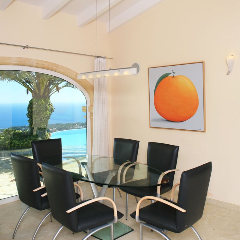 Eetruimte met glazen tafel en moderne stoelen, verrijkt met een grote boogvormige raampartij die panoramisch uitzicht biedt op de zee en kustlijn. Een kunstwerk van een sinaasappel siert de muur.
