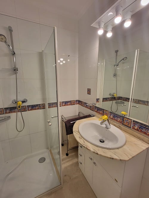 Functionele badkamer met glazen douchecabine, wastafelmeubel met licht marmeren blad en verlichte spiegel. Witte tegels met decoratieve mediterrane rand.
