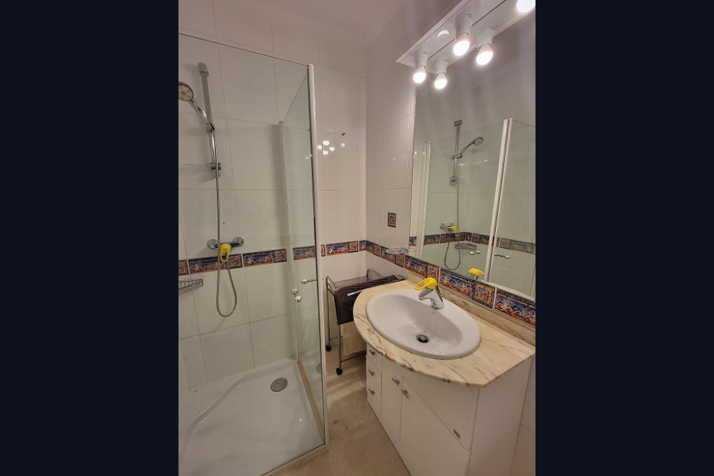 Functionele badkamer met inloopdouche met glazen scherm, wastafel in een licht marmeren blad en wit badmeubel. Witte wandtegels zijn voorzien van een decoratieve mediterrane rand.