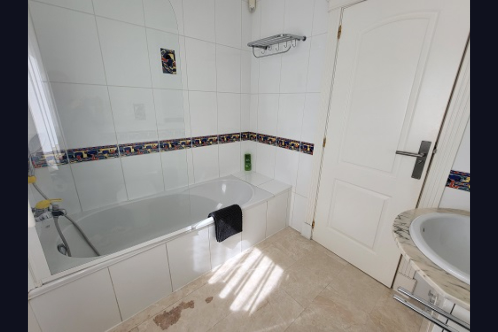 Functionele badkamer met wit ligbad, glazen douchescherm en witte tegelwanden met decoratieve rand. Voorzien van marmeren wastafel en natuurlijk lichtinval.