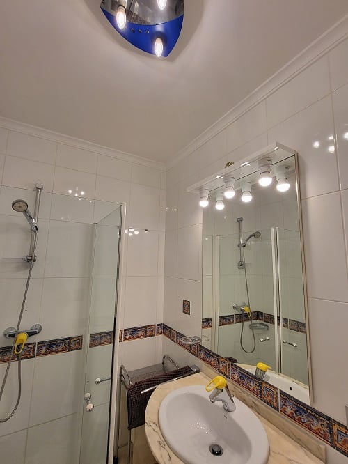 Functionele badkamer met witte tegels en decoratieve rand, glazen douchecabine en wastafel met marmerlook blad. Verlichte spiegel en blauwe plafondlamp.