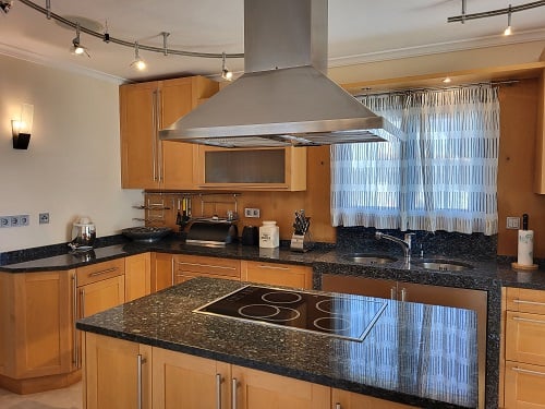 Functionele keuken met lichte houten kasten en donkere granieten werkbladen. Het kookeiland is voorzien van een inductiekookplaat onder een RVS afzuigkap.