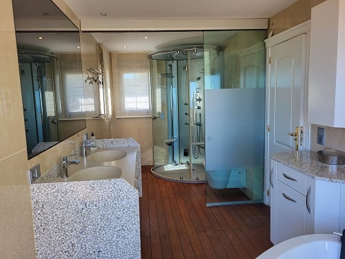 Hoofdbadkamer met dubbele wastafel op unieke kiezelbasis, ruime glazen massagedouche en warme houtlook vloer. Natuurlijk licht via de ramen.