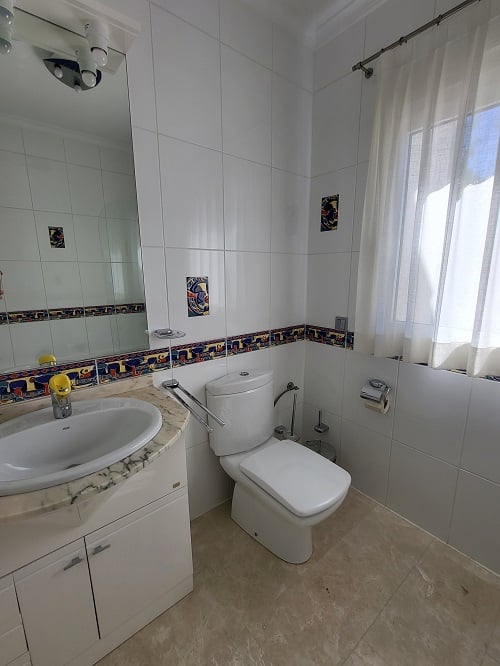 Lichte badkamer met witte wandtegels, een kleurrijke decoratieve rand en accentstukken. Voorzien van een marmeren wastafelmeubel met geïntegreerde wasbak, een modern toilet en een raam met vitrage.