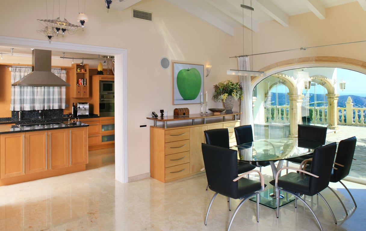 Lichte eetruimte met glazen tafel en zwarte stoelen, op gepolijste marmeren vloeren. Grote boogvensters bieden panoramisch zee- en bergzicht, grenzend aan een moderne keuken.
