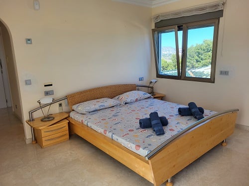 Lichte slaapkamer met een licht houten bed, rieten hoofdeinde en nachtkastje. Het open raam biedt uitzicht op weelderig groen en verre bergen.