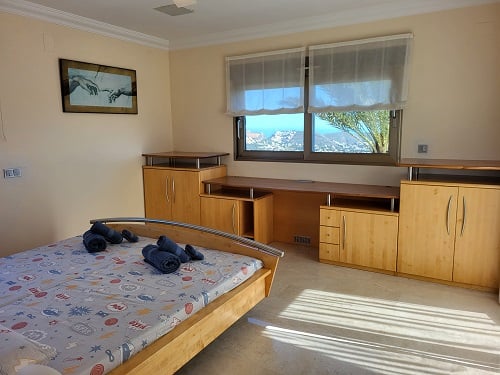 Lichte slaapkamer met licht houten bed, geïntegreerd bureau en kasten. De ramen bieden panoramisch uitzicht op de kust, waardoor de ruimte wordt gevuld met natuurlijk licht.
