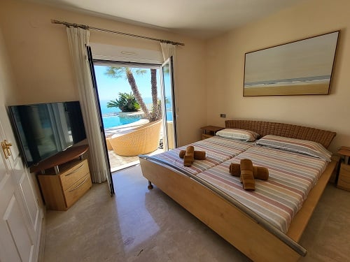 Lichte slaapkamer met twee eenpersoonsbedden en gevlochten hoofdbord, directe toegang tot het zwembad en uitzicht op zee. Inclusief televisie en muurkunst.