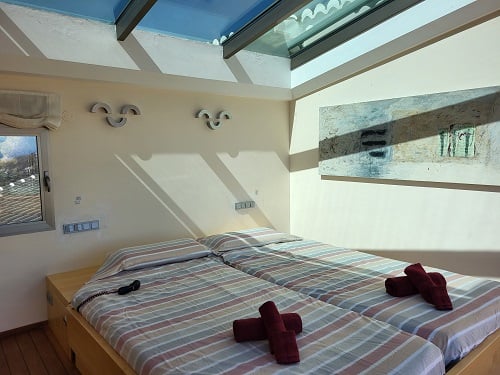 Lichte slaapkamer met twee eenpersoonsbedden, gestreept beddengoed en rode handdoeken. Een groot dakraam voorziet de kamer van natuurlijk licht, aangevuld met een abstract schilderij.