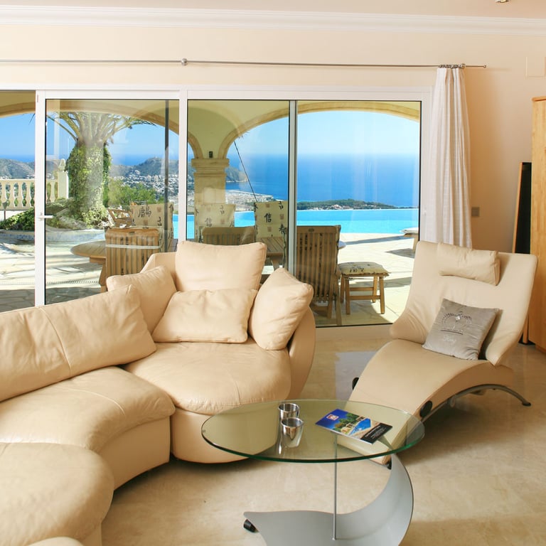 Lichte woonkamer met crèmekleurige, gebogen banken en marmeren vloer, direct verbonden met een terras met overloopzwembad en panoramisch uitzicht op zee.