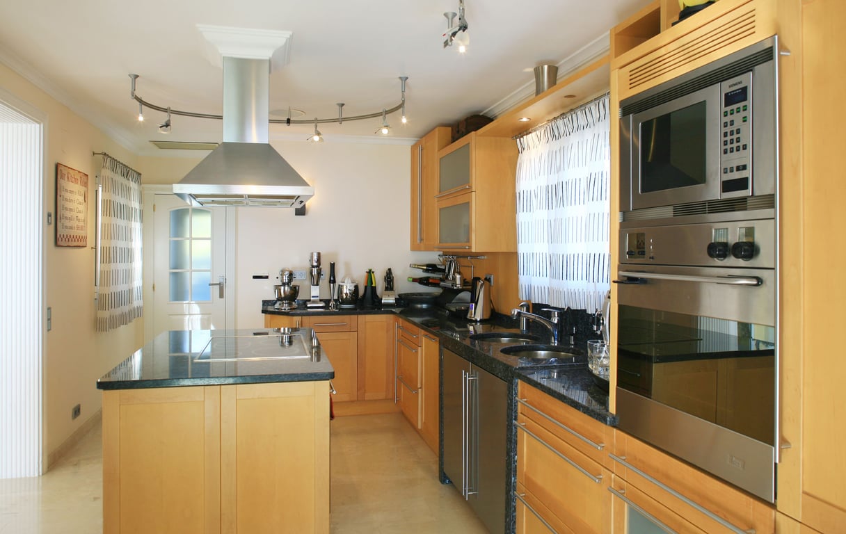 Moderne keuken met lichte houten kasten, donkere granieten werkbladen en een kookeiland met geïntegreerde kookplaat. Roestvrijstalen apparatuur omvat oven, magnetron en afzuigkap.