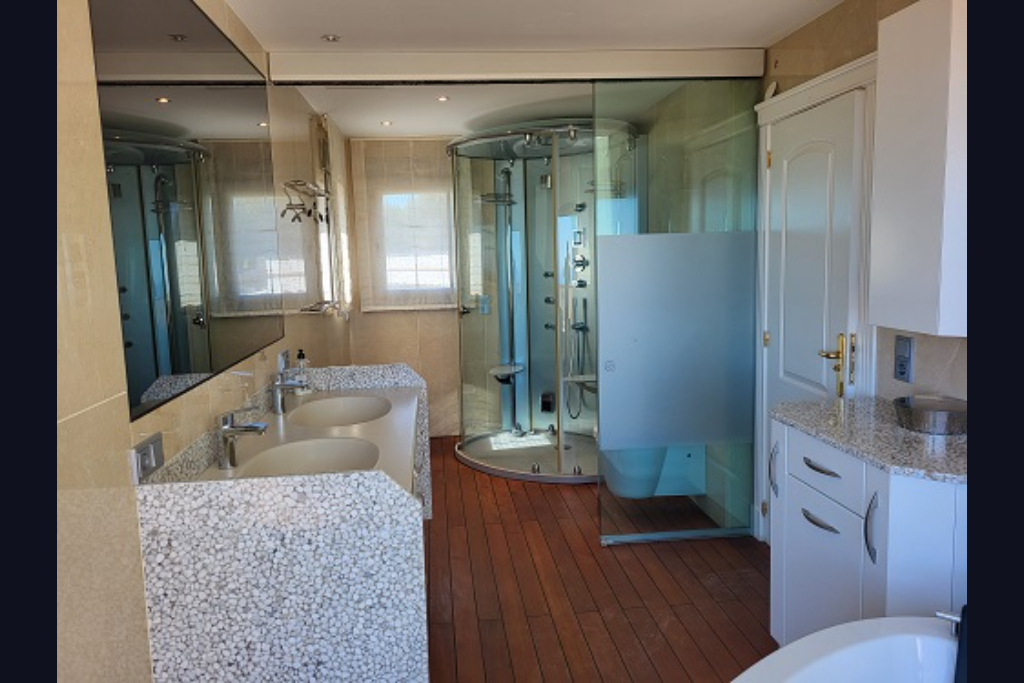 Ruime badkamer met dubbele wastafel in kiezelsteen-design, ronde hydromassage douche en warme houten vloer. Grote spiegel en ramen met jaloezieën aanwezig.
