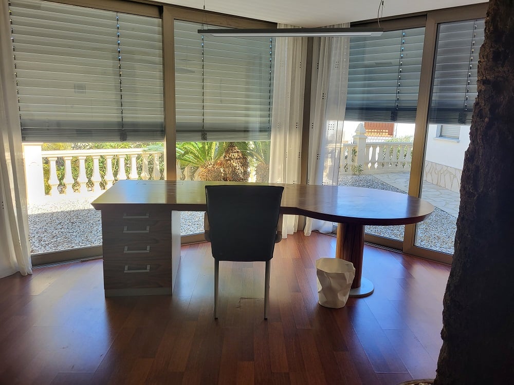 Ruime privékantoor met op maat gemaakt houten bureau en kamerhoge ramen. Uitzicht op grindterras en mediterrane beplanting. Donkere parketvloer en moderne verlichting.