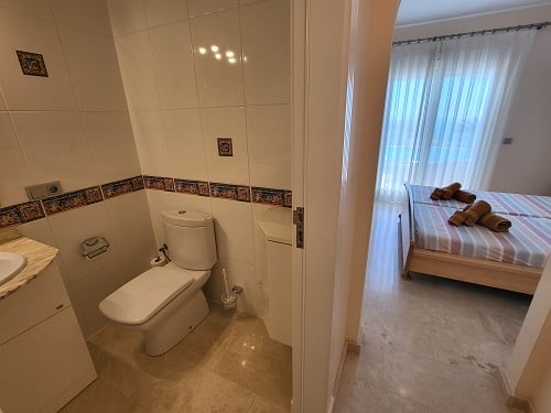 Zicht vanuit een betegelde badkamer met toilet en wastafel, naar een lichte slaapkamer met eenpersoonsbedden, gestreept beddengoed en groot raam.
