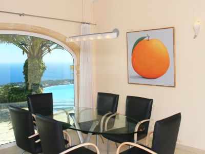 Eetruimte met glazen tafel en moderne stoelen, verrijkt met een grote boogvormige raampartij die panoramisch uitzicht biedt op de zee en kustlijn. Een kunstwerk van een sinaasappel siert de muur.