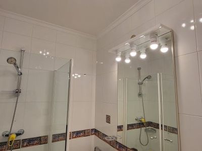 Functionele badkamer met witte tegels en decoratieve rand, glazen douchecabine en wastafel met marmerlook blad. Verlichte spiegel en blauwe plafondlamp.