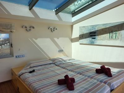 Lichte slaapkamer met twee eenpersoonsbedden, gestreept beddengoed en rode handdoeken. Een groot dakraam voorziet de kamer van natuurlijk licht, aangevuld met een abstract schilderij.