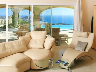 Lichte woonkamer met crèmekleurige, gebogen banken en marmeren vloer, direct verbonden met een terras met overloopzwembad en panoramisch uitzicht op zee.