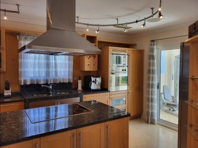 Moderne keuken met lichte houten kasten, donkere granieten werkbladen, een kookeiland met inductiekookplaat en roestvrijstalen apparatuur. Glazen deur naar fitnessruimte.