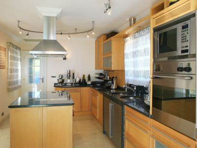 Moderne keuken met lichte houten kasten, donkere granieten werkbladen en een kookeiland met geïntegreerde kookplaat. Roestvrijstalen apparatuur omvat oven, magnetron en afzuigkap.
