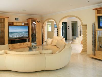 Ruime woonkamer met een grote crèmekleurige gebogen bank, lichte marmeren vloeren en op maat gemaakte lichte houten tv-meubels. Stenen bogen leiden naar de hal en trap.