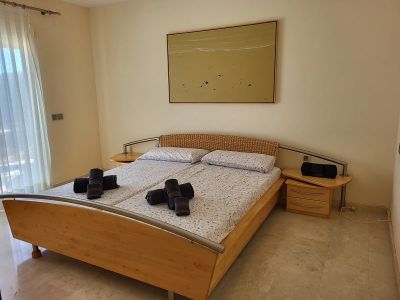 Slaapkamer met houten bedframe, gevlochten hoofdeinde en metalen accenten. Nachtkastjes met lades flankeren het bed, onder een abstract schilderij. Natuurlijk licht valt binnen via vitrage.