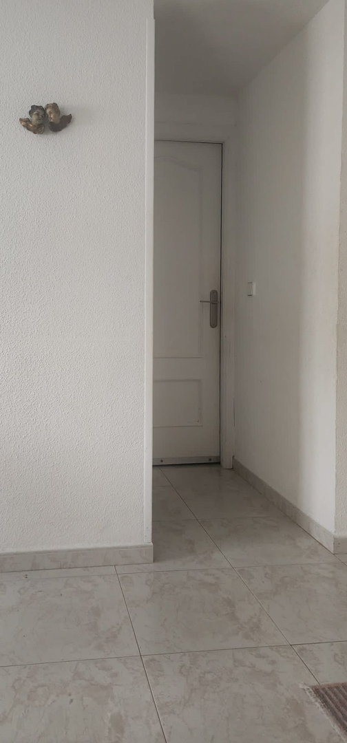 Binnengang met lichte tegelvloer en wit gestucte muren, leidend naar een paneeldeur, met een klein decoratief wanddetail.