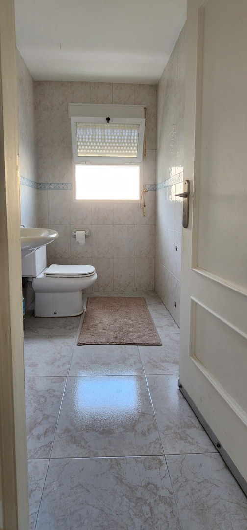 Compacte badkamer met toilet, wandgemonteerde wastafel en raam met zonwering. Lichte tegelwanden en vloer aanwezig.