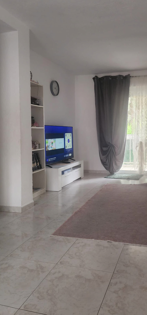 De woonkamer heeft lichte tegelvloeren en witte muren, met een modern tv-meubel en een boekenkast. Natuurlijk licht valt binnen via de toegang tot het terras, omkaderd door donkergrijze en witte gordijnen.