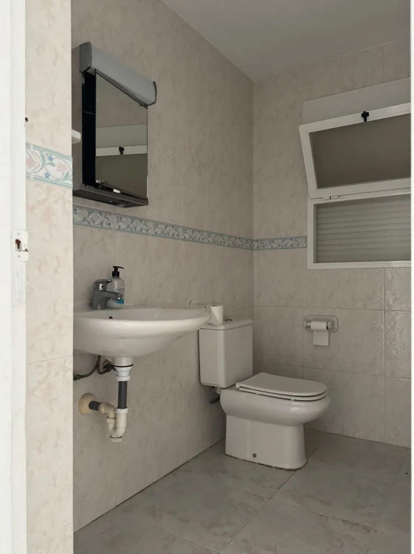 Functionele badkamer met lichte tegels, decoratieve rand en een kiepraam. Voorzien van een hangende wastafel, toilet en spiegelkast met geïntegreerde verlichting.