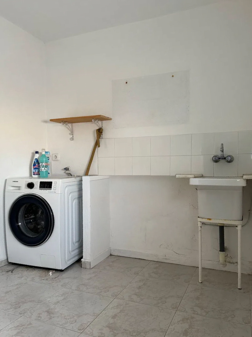 Functionele bijkeuken met een moderne wasmachine, een eenvoudige spoelbak en witte tegelwand. Voorzien van lichte vloertegels en een simpele houten plank.