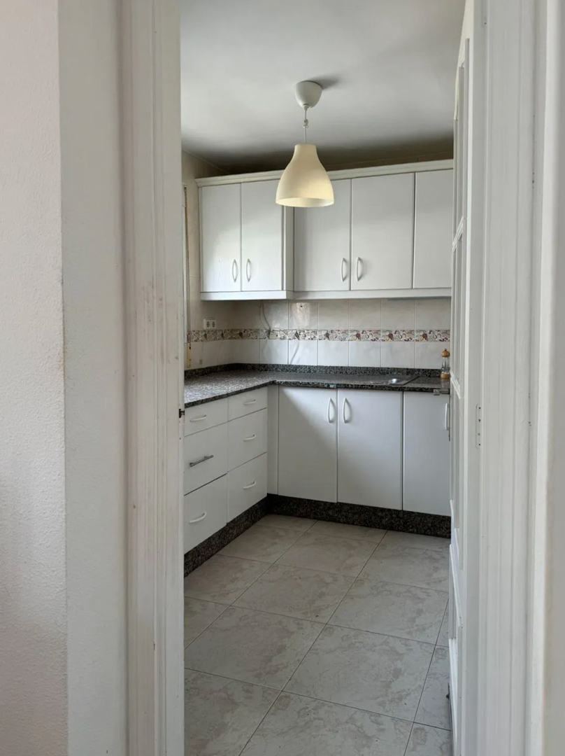 Functionele keuken met witte kasten, een donker gespikkeld granieten aanrechtblad en betegelde achterwand. Een hanglamp verlicht de ruimte, klaar voor modernisering.