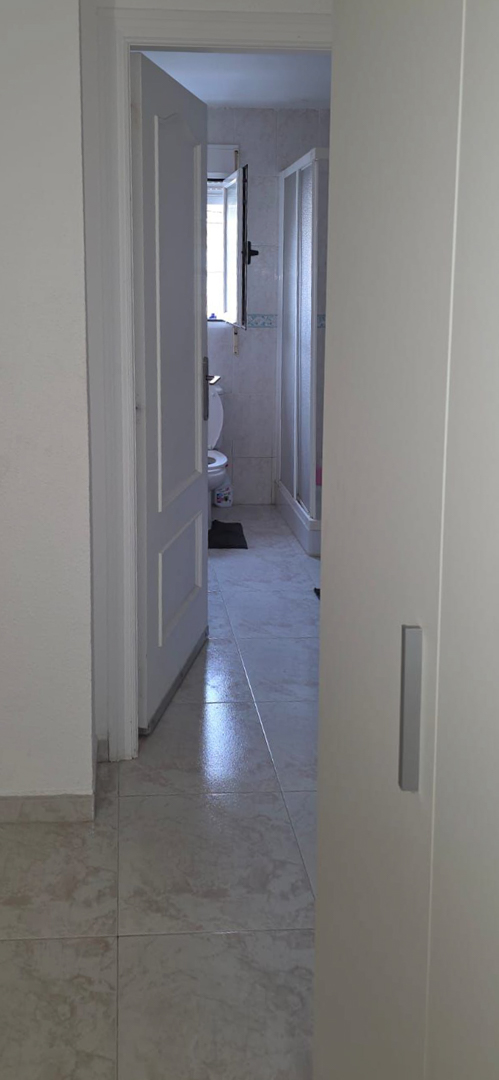 Gedeeltelijk zicht op een badkamer met lichte tegelvloer, toilet, raam voor natuurlijk licht en douchecabine. De wandtegels hebben een decoratieve rand.