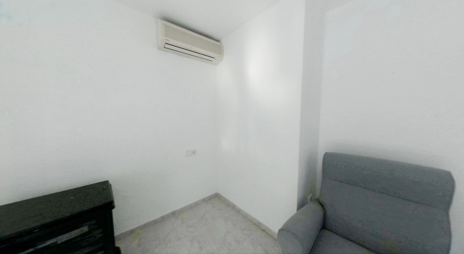 Hoek van een kamer met witte muren en lichte tegelvloer. Een split-airconditioner is geïnstalleerd, met een stopcontact eronder, naast een grijze fauteuil en donker meubelstuk.