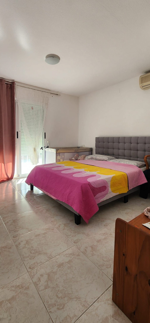 Slaapkamer met grijs gestoffeerd bed, lichte tegelvloer, natuurlijk licht van raam met rolluik en een babybedje. Voorzien van wandairconditioning.