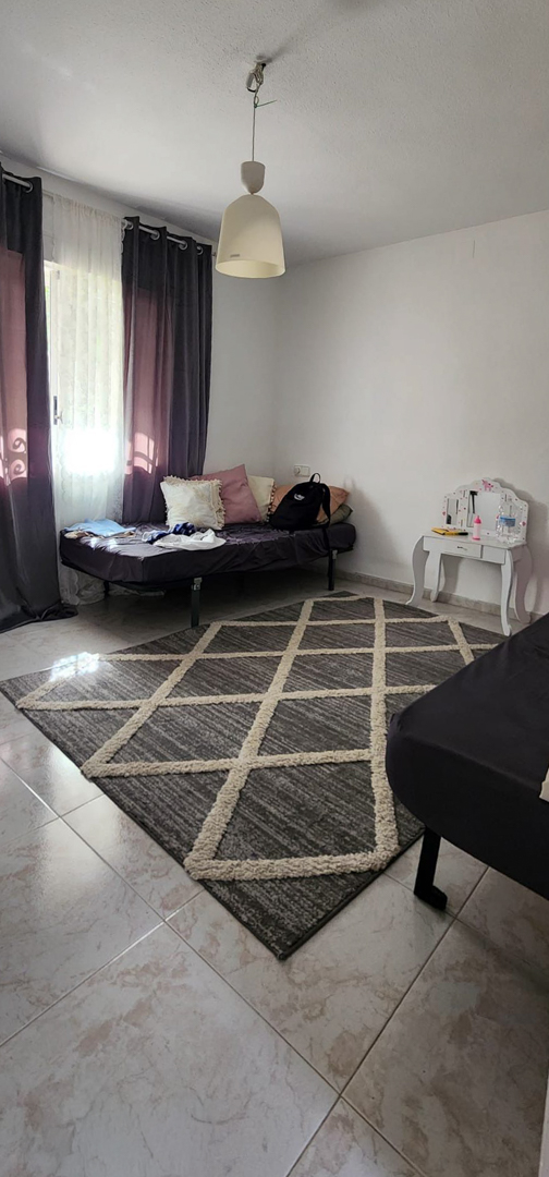 Slaapkamer met lichte tegelvloer, een grijs-beige geometrisch vloerkleed en een eenpersoonsbed bij het raam met donkere gordijnen. Een kleine witte make-uptafel is aanwezig.