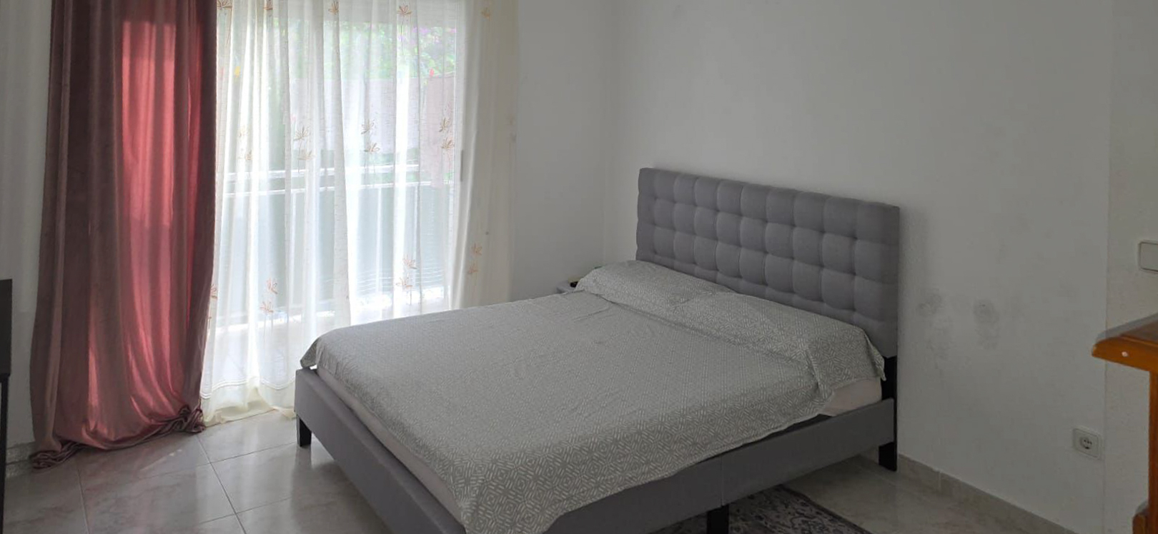Tweepersoons slaapkamer met grijs gestoffeerd bed en gecapitonneerd hoofdbord, tegelvloer en veel natuurlijk licht van het raam met terras toegang.
