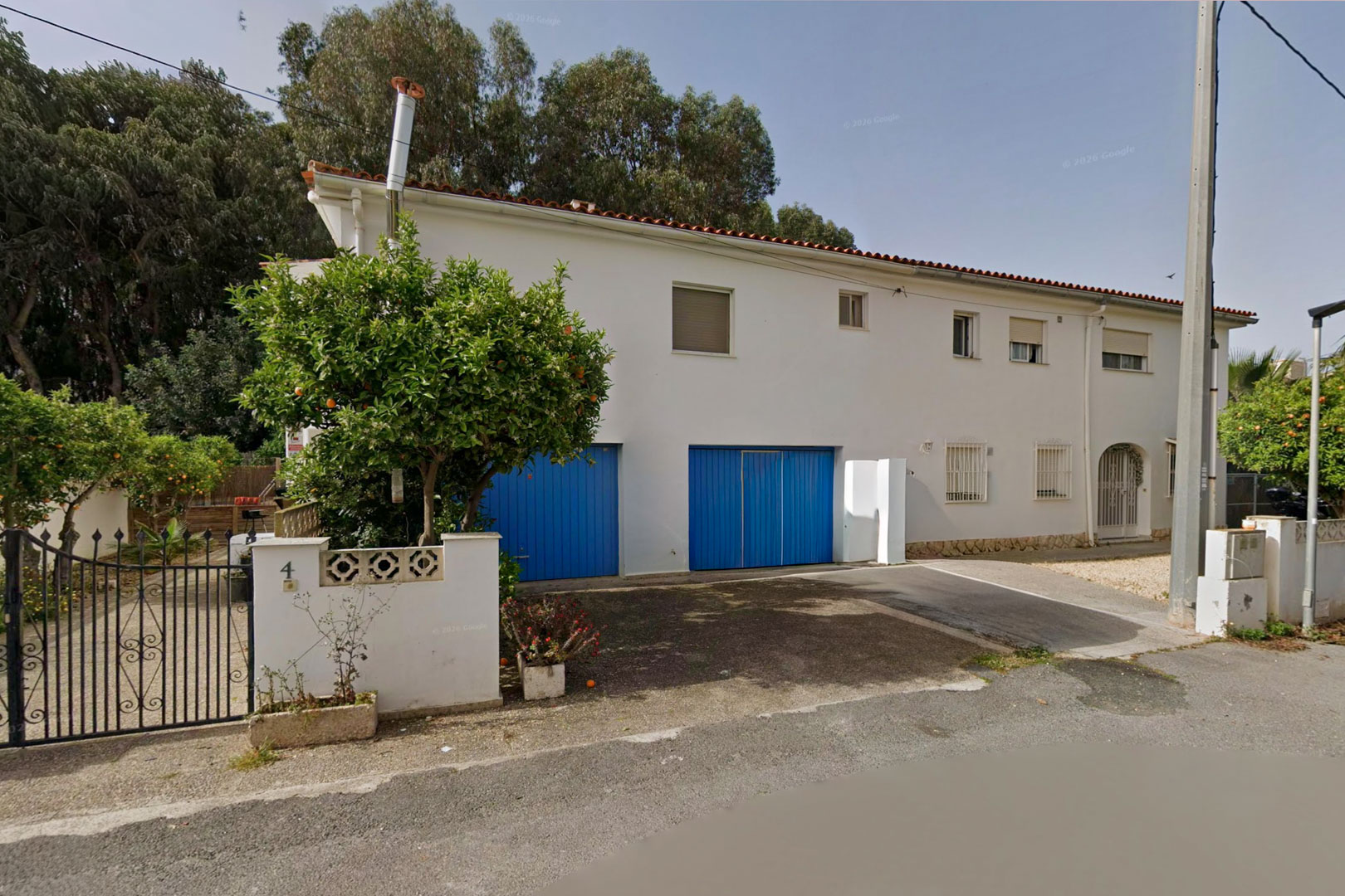 Voorgevel van de halfvrijstaande villa, met een witte gevel, twee opvallende blauwe garagedeuren en een sinaasappelboom bij het smeedijzeren toegangshek.