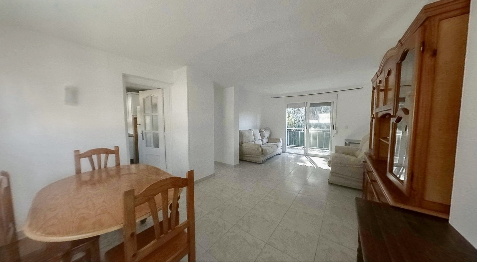 Zicht op de lichte woon-eetkamer met een ovale houten tafel, tegelvloeren en een grote houten vitrinekast. Glazen schuifdeuren openen naar een balkon, waardoor de ruimte natuurlijk licht ontvangt.