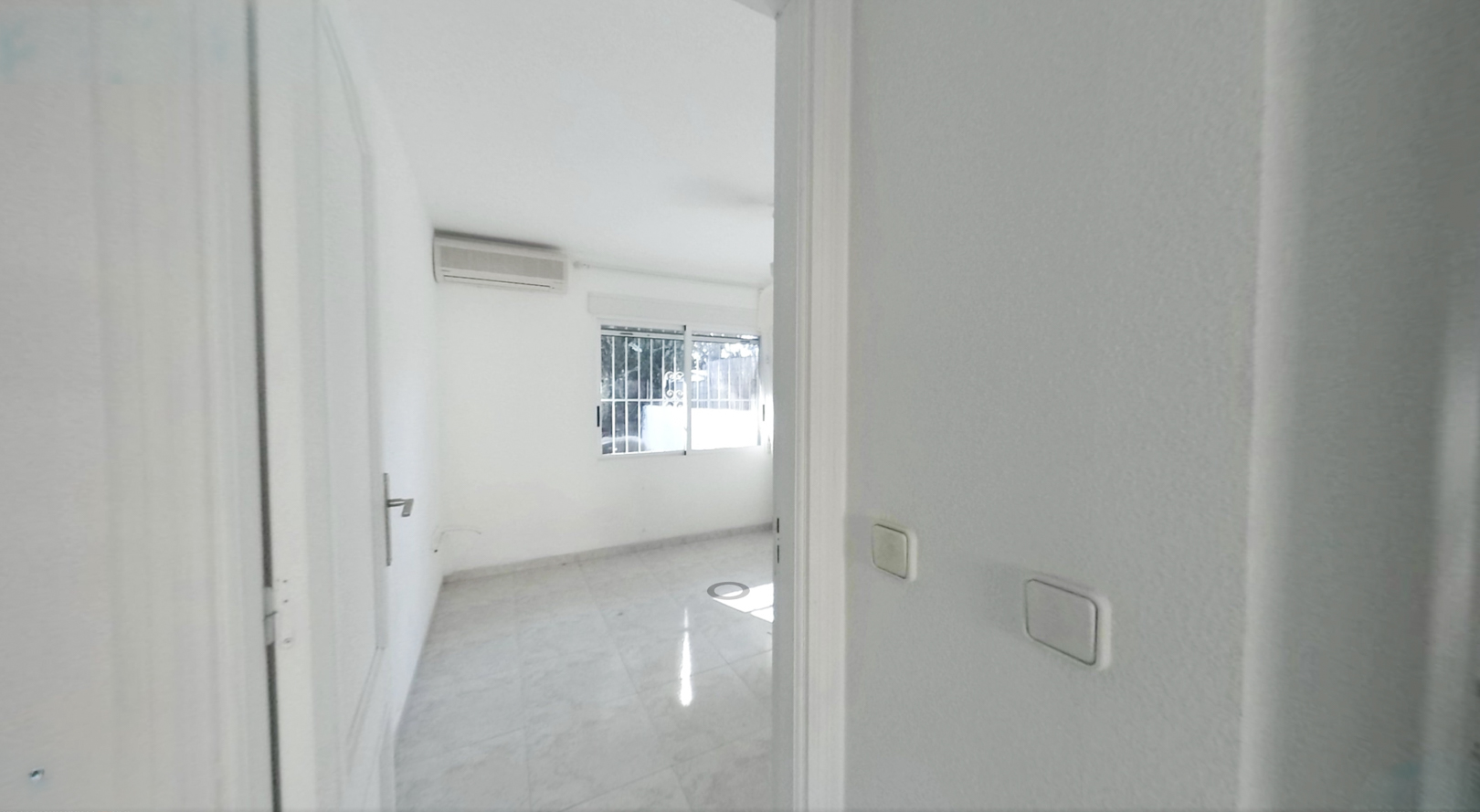 Zicht op een lege kamer met lichte tegelvloer, witte muren en een airconditioningunit. Het raam met tralies zorgt voor natuurlijk daglicht.