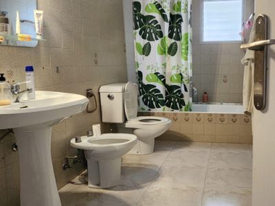 Badkamer met lichtgekleurde tegels, een vrijstaande wastafel, toilet, bidet en ligbad met tropische douchegordijn en matglazen raam.