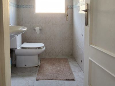Compacte badkamer met toilet, wandgemonteerde wastafel en raam met zonwering. Lichte tegelwanden en vloer aanwezig.