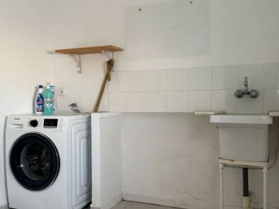 Functionele bijkeuken met een moderne wasmachine, een eenvoudige spoelbak en witte tegelwand. Voorzien van lichte vloertegels en een simpele houten plank.