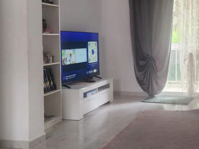 Gedeeltelijk zicht op de woonkamer met televisie op een wit meubel, een rek en toegang tot een terras met grijze en transparante gordijnen, op een lichte tegelvloer met vloerkleed.