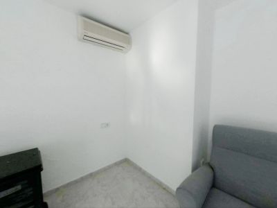 Hoek van een kamer met witte muren en lichte tegelvloer. Een split-airconditioner is geïnstalleerd, met een stopcontact eronder, naast een grijze fauteuil en donker meubelstuk.