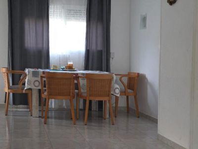 Lichte eethoek met houten tafel en gevlochten stoelen, geplaatst bij een raam met grijze gordijnen. De vloer is licht betegeld, met een gestreept vloerkleed op de voorgrond.
