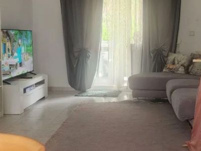 Ruime woonkamer met natuurlijk licht, voorzien van tegelvloeren, een comfortabele hoekbank en directe toegang tot een terras via glazen deuren met gordijnen.