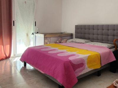 Slaapkamer met grijs gestoffeerd bed, lichte tegelvloer, natuurlijk licht van raam met rolluik en een babybedje. Voorzien van wandairconditioning.